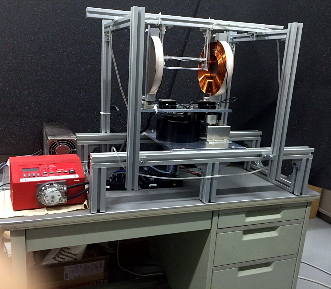 The redesigned electromagnetic coil pair on its aluminum test bench, bare copper spiral visible, with a red pump simulating blood flow