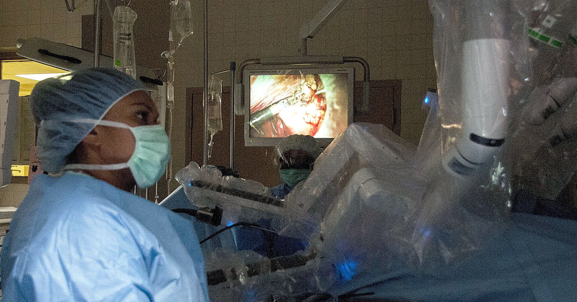 A masked surgical nurse watches the video feed beside the draped arms of a da Vinci robot during an operation