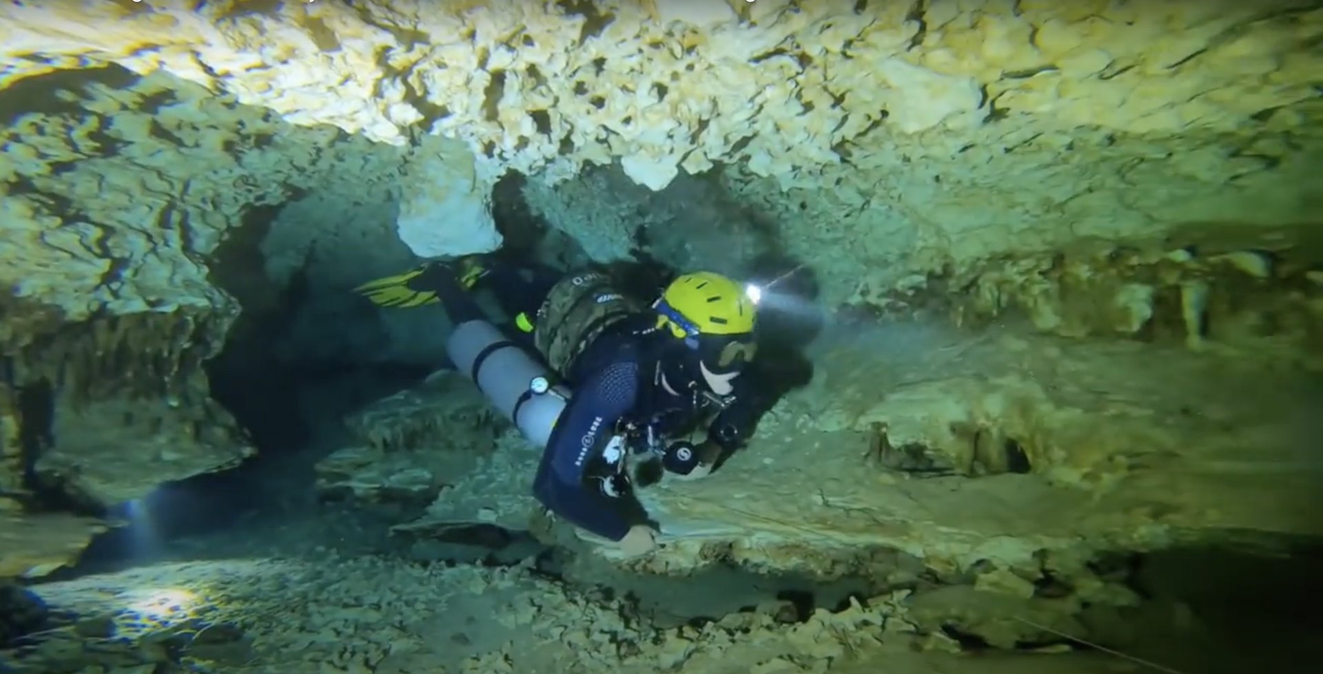 A cave diver deep inside a flooded cave, lit only by their own helmet light, rock on every side and no daylight anywhere