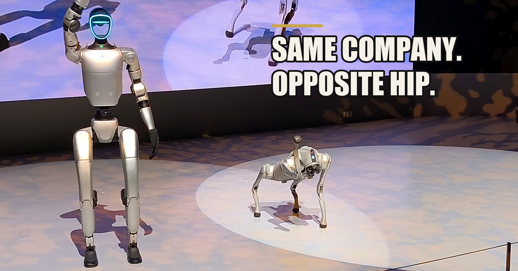 A silver Unitree G1 humanoid robot on a show stage with one hand raised, its legs and hip actuators visible, a four-legged robot dog beside it